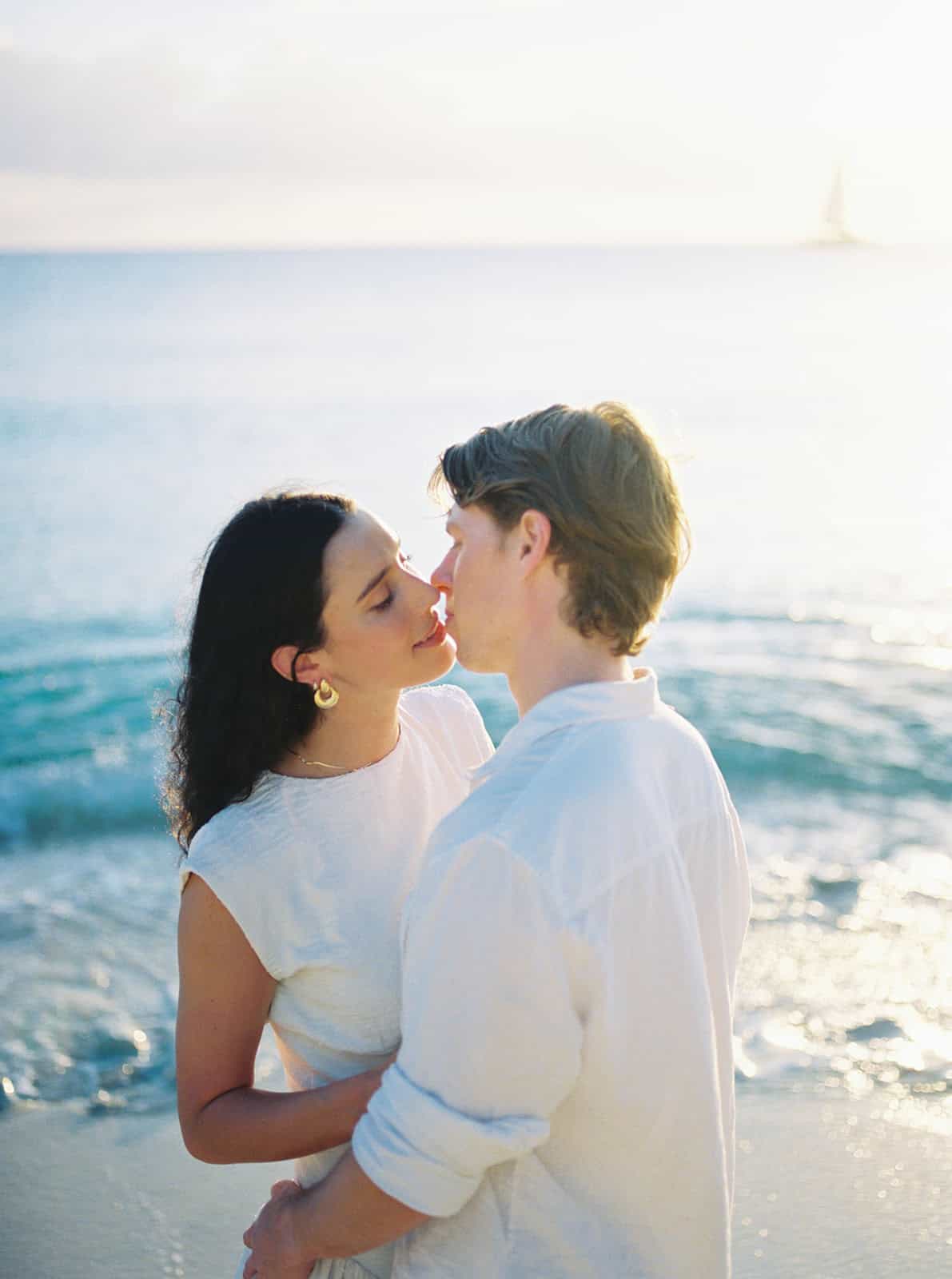 Couple sharing a kiss at the water's edge with a sailboat behind them during their grand cayman engagement session